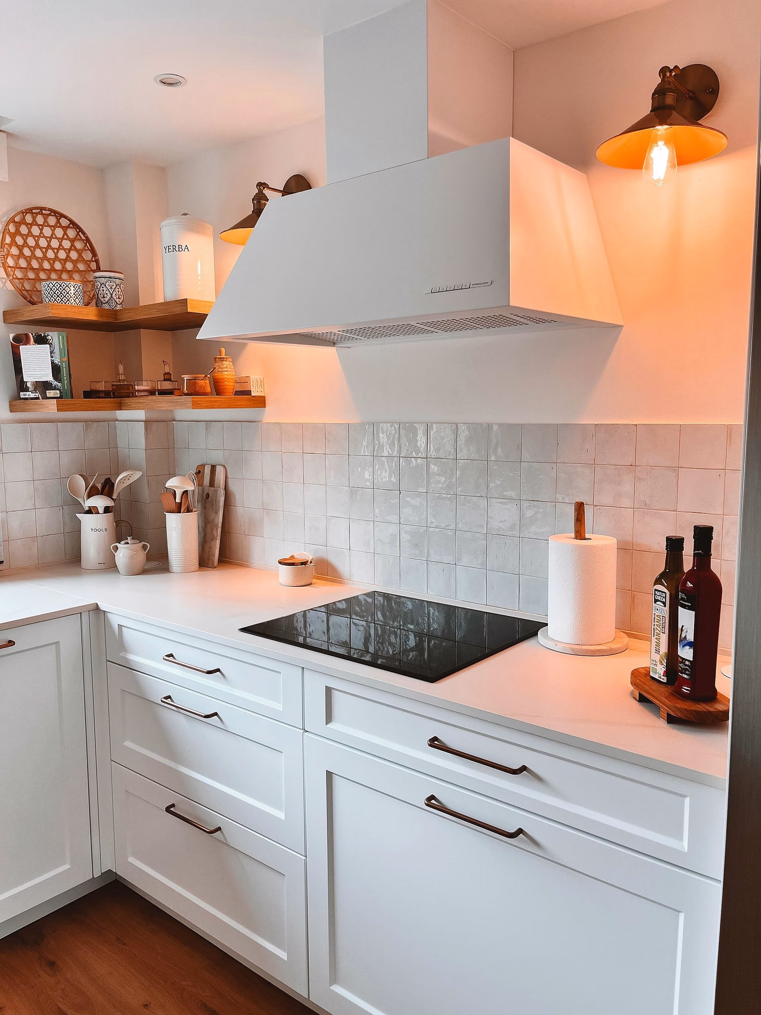 Cocina en L blanca con campana decorativa, encimera clara y baldas de madera.